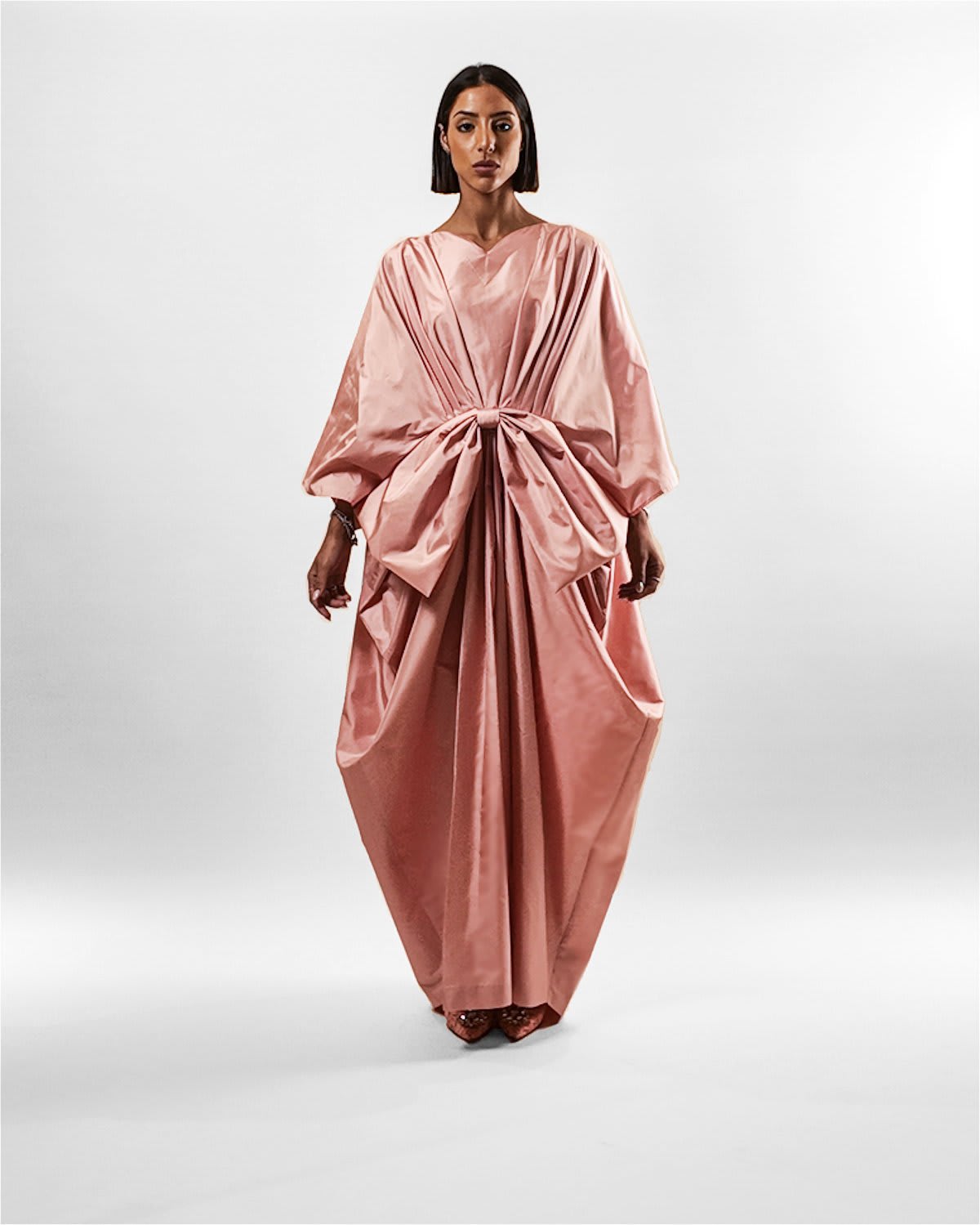 rosetta silk bow kaftan Tanisha Rada model wearing the in Rosetta Silk Bow Kaftan, featuring dramatic batwing sleeves and a massive structural bow gathered at the waist, creating a voluminous, draped silhouette against a neutral beige background. front