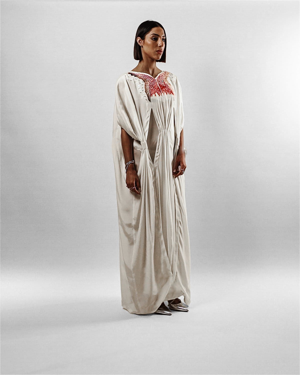 cocora kaftan A full-length studio portrait of a woman with a sleek, dark bob haircut wearing the Cocora Kaftan by Tanisha Rada. The floor-length garment is crafted from a fluid, cream-colored silk with an intricate red and gold butterfly-shaped embroidery across the chest. It features draped, cape-like sleeves and unique geometric fabric overlays that create a structured, multi-dimensional silhouette against a neutral beige background. side