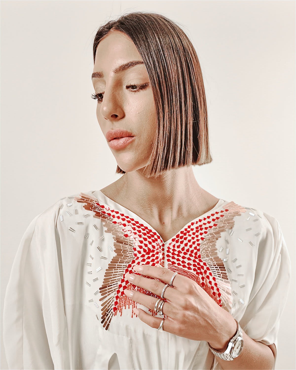 cocora kaftan A full-length studio portrait of a woman with a sleek, dark bob haircut wearing the Cocora Kaftan by Tanisha Rada. The floor-length garment is crafted from a fluid, cream-colored silk with an intricate red and gold butterfly-shaped embroidery across the chest. It features draped, cape-like sleeves and unique geometric fabric overlays that create a structured, multi-dimensional silhouette against a neutral beige background. close up