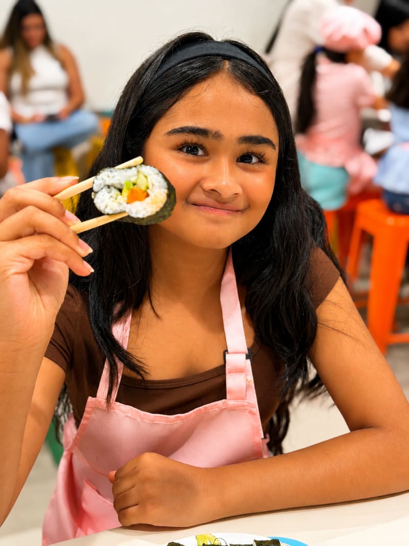 A child in an apron holding a sushi roll she rolled during CocinArte