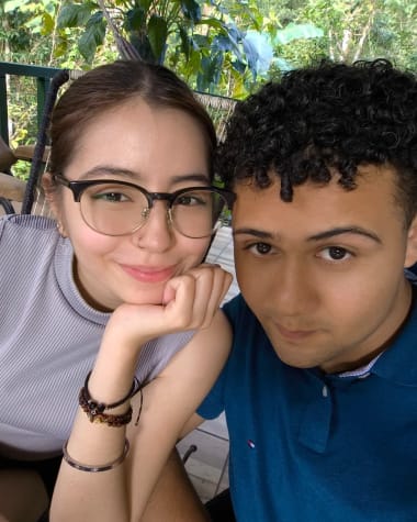 Katheryn apoyando la barbilla en la mano, con Brandon a su lado y vegetacion al fondo