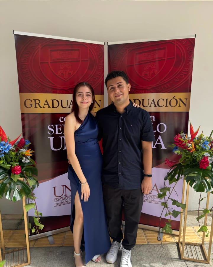Katheryn y Brandon abrazados frente a los banners de la graduacion de la Universidad Nacional