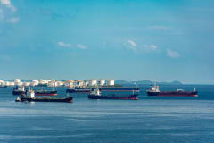 A fleet of cargo ships docked near oil storage tanks along a serene coastline with a clear blue sky above.