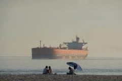 Oil tanker anchored near a beach where people relax under an umbrella.