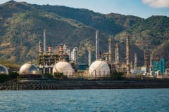 Industrial refinery with spherical tanks by the coastline against a backdrop of mountains.