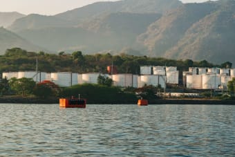 Oil storage tanks located by a calm waterway, with a mountainous backdrop under a clear sky.