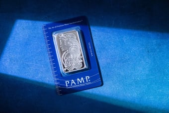Silver bar in PAMP packaging on a textured blue surface with sunlight.