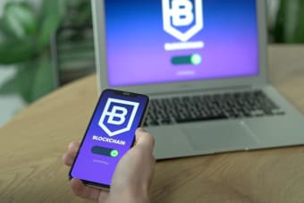 A hand holding a smartphone displaying a blockchain app, with a laptop on a desk in the background.