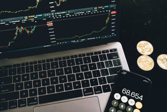 A laptop with trading charts, smartphone calculator, and bitcoin coins depicting cryptocurrency trading.