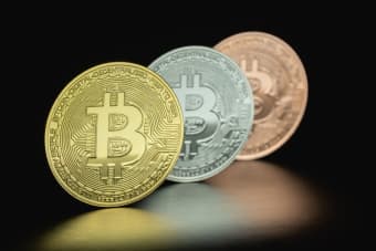 Close-up image of three Bitcoin coins in gold, silver, and bronze tones against a dark background.