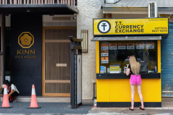 Woman at currency exchange booth in Pattaya, Thailand, with currency rates displayed.