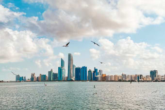 Stunning view of Abu Dhabi skyline with birds flying above the sea under a cloudy sky.