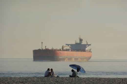 Oil tanker anchored near a beach where people relax under an umbrella.