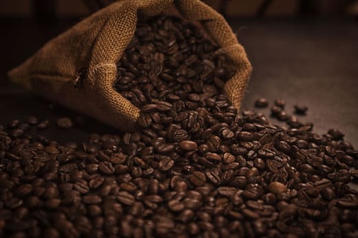 A burlap sack tipping over with dark roasted coffee beans spreading on a surface.