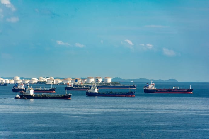A fleet of cargo ships docked near oil storage tanks along a serene coastline with a clear blue sky above.