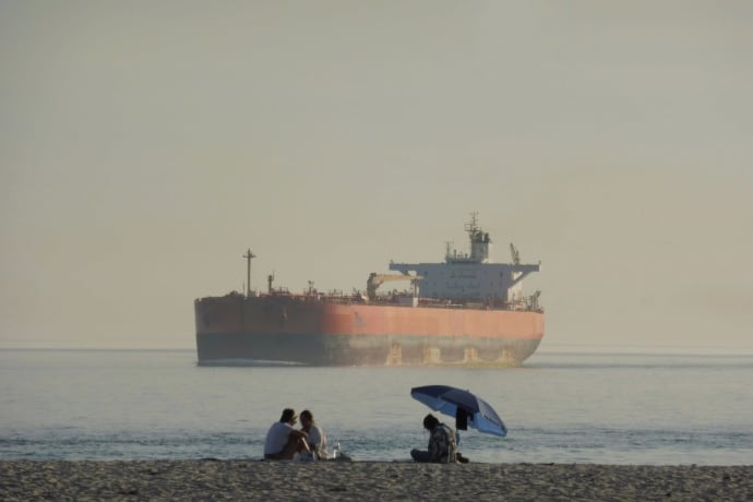 Oil tanker anchored near a beach where people relax under an umbrella.