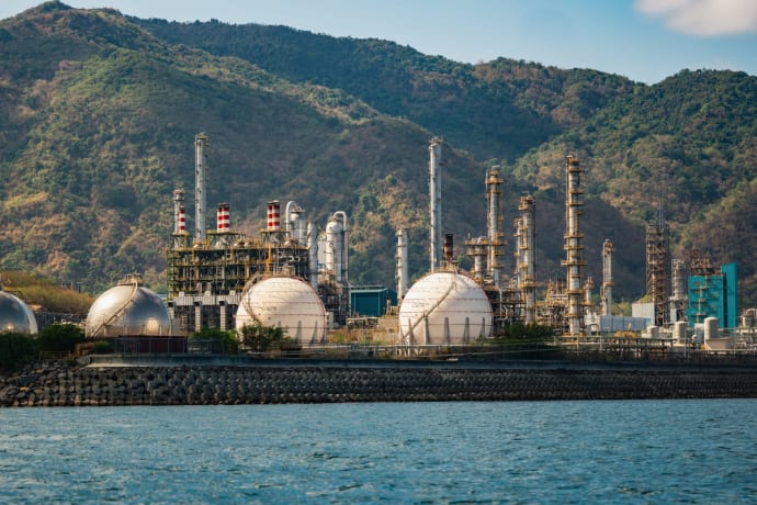 Industrial refinery with spherical tanks by the coastline against a backdrop of mountains.