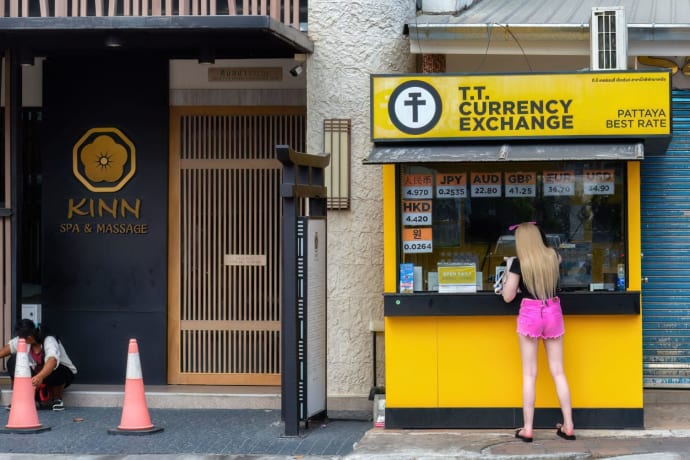 Woman at currency exchange booth in Pattaya, Thailand, with currency rates displayed.
