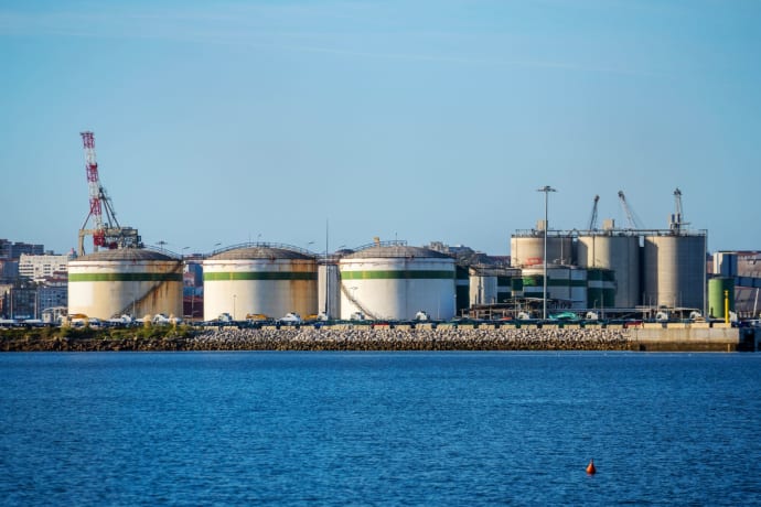 Large storage tanks at an industrial port facility by the water under clear blue skies.