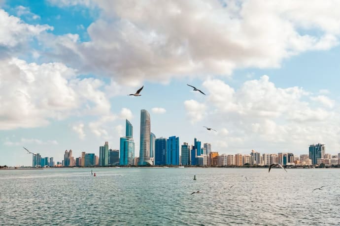 Stunning view of Abu Dhabi skyline with birds flying above the sea under a cloudy sky.