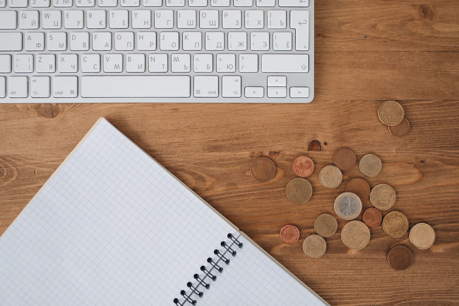 Flat lay of a desk featuring a keyboard, notebook, and scattered coins on a wooden surface.