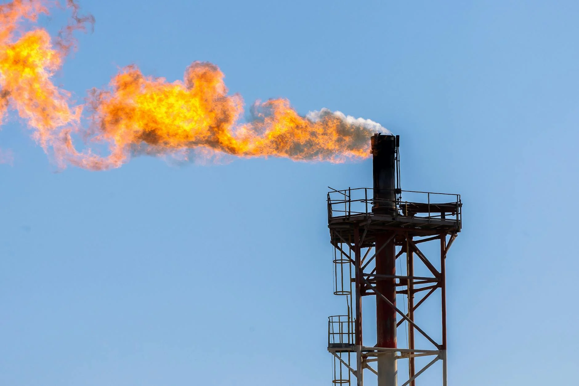 Close-up of an industrial gas flare with a vibrant orange flame against a clear blue sky.