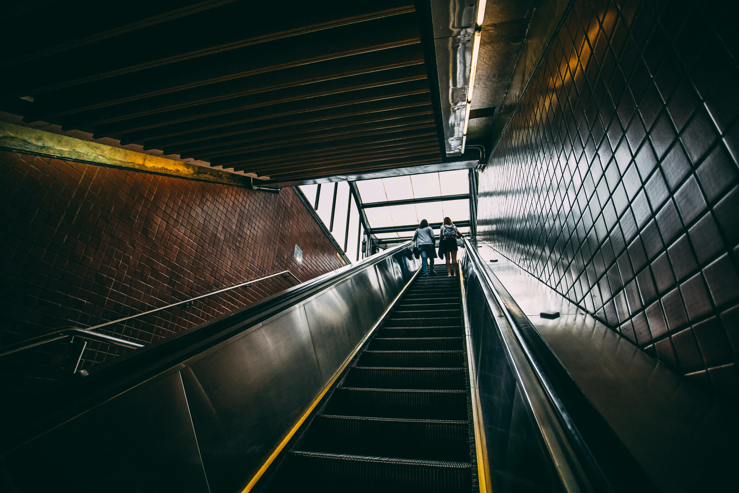 Two women ascend on an indoor escalator in a modern urban setting.