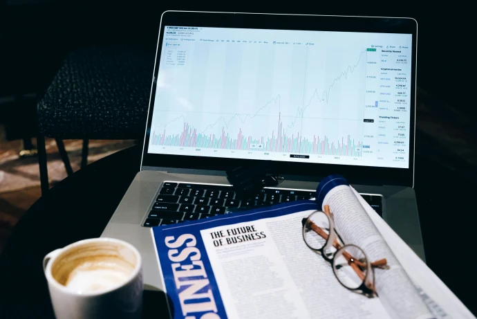A modern business workspace featuring a laptop showing stock data next to a business magazine and coffee.
