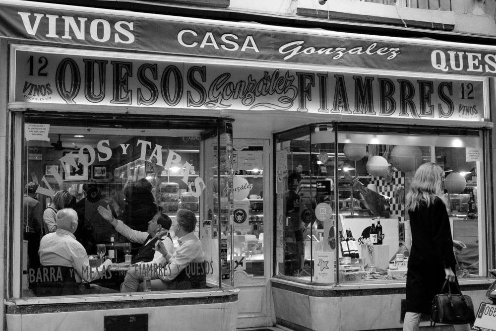 Men discussing and gesticulating in a Spanish bar while woman walks by outside.