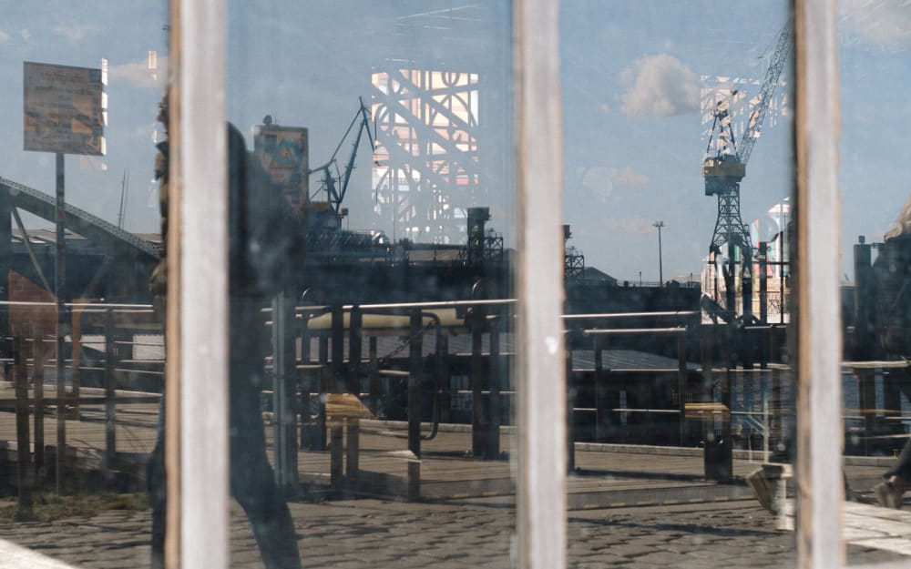 Window reflection showing harbour cranes, patterns from the window decorations, and somewhat visible people.