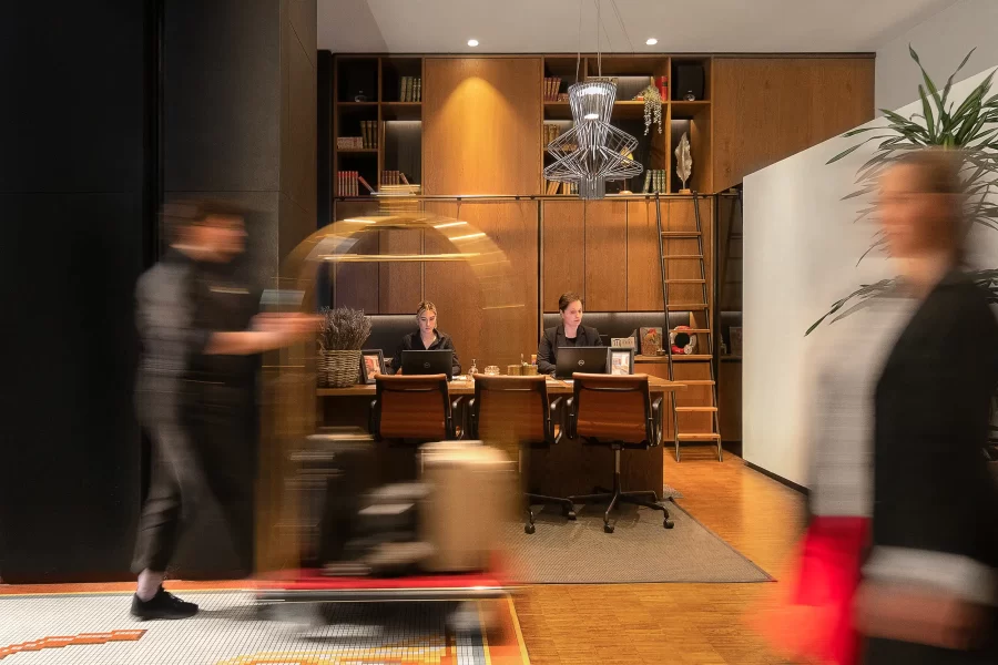 Hotelfotografie der Lobby des Mosaic House in Prag mit bewegungsunscharfem Portier und Gästen, wobei der Gepäckwagen eine natürliche Rahmung der Rezeption bildet.