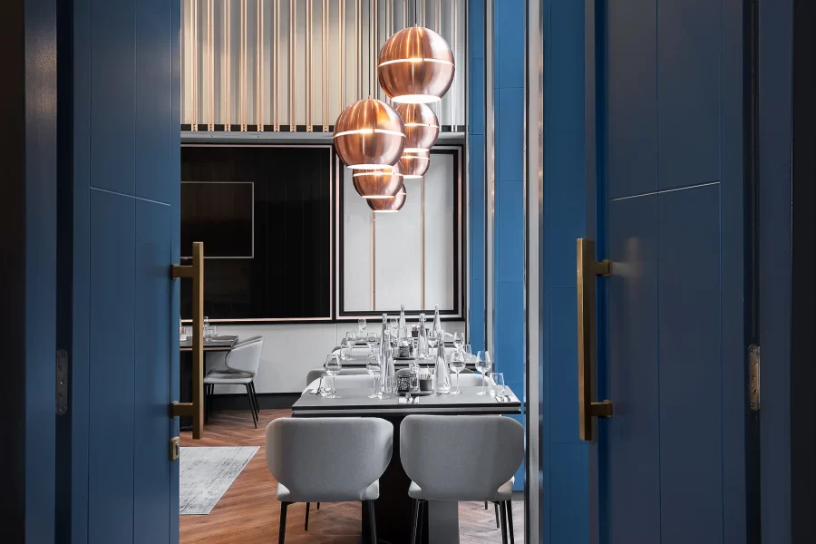 Průhled pootevřenými dveřmi na prostřené stoly v útulné a elegantní restauraci v hotelovém baru Radisson Blu v Praze, odrážející jeho pohostinnost. Restaurace je vyzdobena modro-bronzovými barvami.
