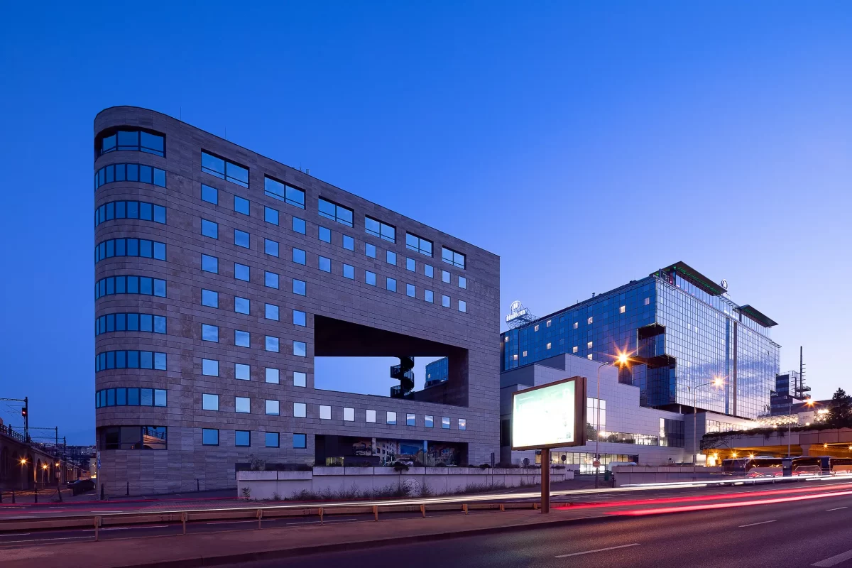 A photograph of the International Business Centre (IBC) in Prague's Karlin was taken during the blue hour, showcasing its contextual architecture alongside the Hilton hotel and viaduct.