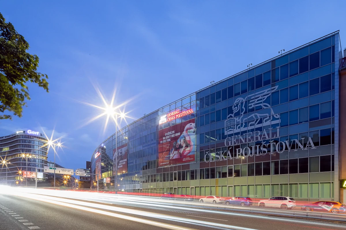 Blue hour overview of Prague Generali headquarters wrapped with continuous cover glass wall with lettering and tinted glass from the 5. Kvetna Street, with light trails from cars passing by, is incorporated into the scene.
