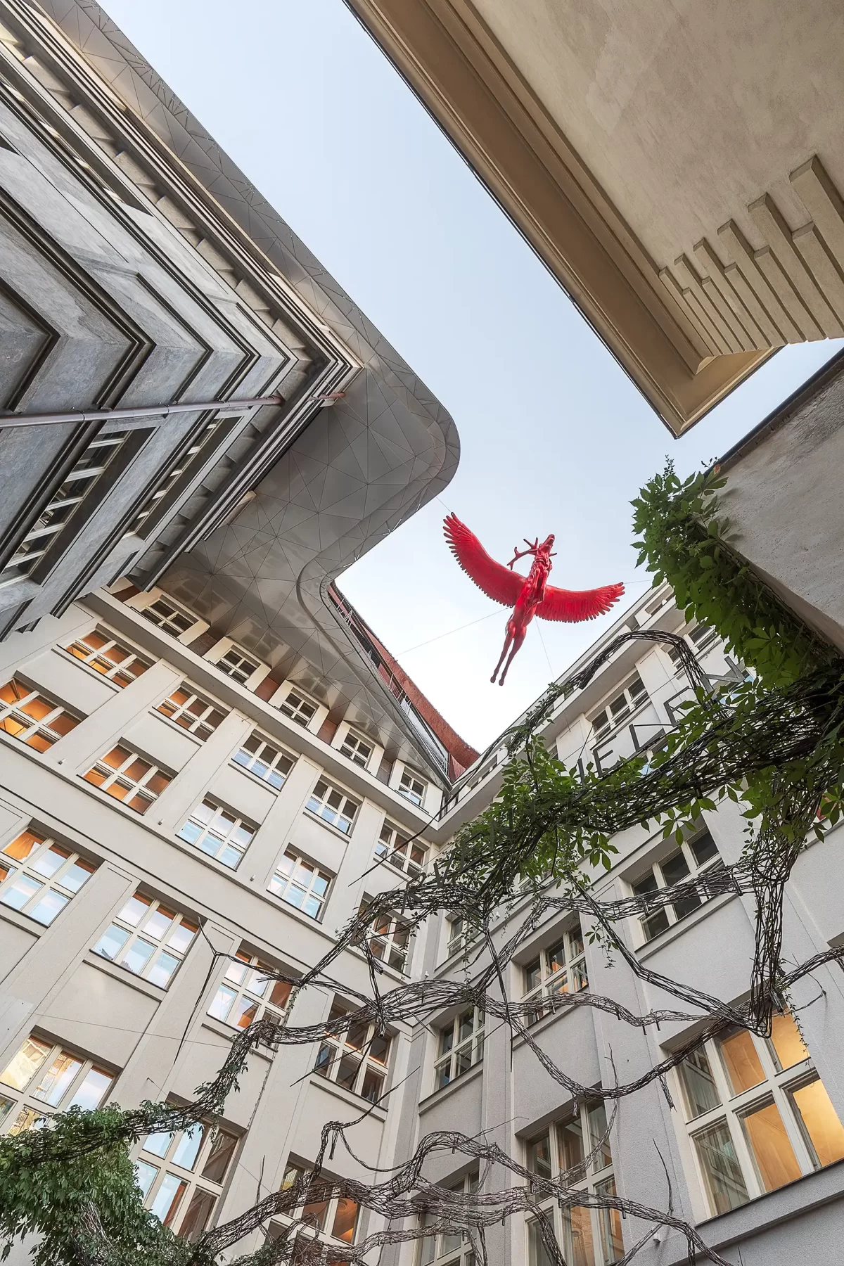 Pohled směrem vzhůru, vyfotografovaný z nádvoří (vnitrobloku) Šporkova paláce v Praze na zavěšenou sochu červeného jelena, umístěnou doprostřed kompozice/průhledu.