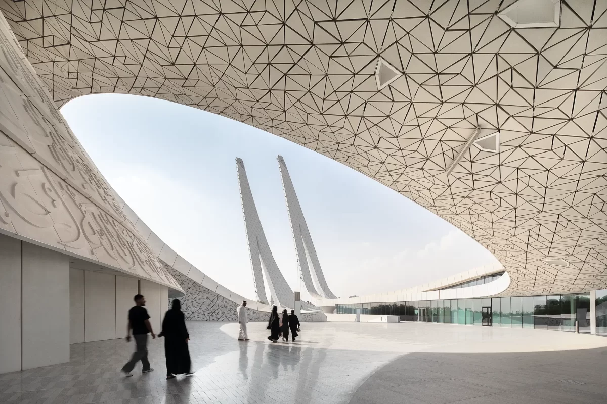 Blick auf die modernen Minaretttürme, eingerahmt vom ovalen Dach des HBKU-Minaretein-Gebäudes in der Education City in Doha (Katar), die sich im Marmorboden spiegeln. Eine Gruppe von Gläubigen, durch die Bewegung verschwommen, verlässt gerade die Moschee.