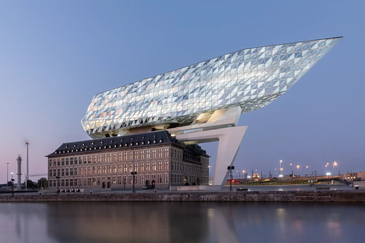 Gesamtansicht des Port House von Zaha Hadid, das sich bei Einbruch der Dämmerung über dem Wasser erhebt – moderne Architektur, die ein historisches Gebäude mit einem futuristischen Anbau im Hafen von Antwerpen verbindet.