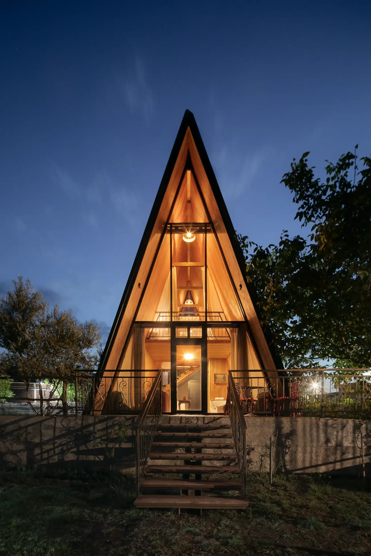 Noční fotografie moderní dvoupatrové dřevostavby glampingového apartmánu se sešikmenou střechou do tvaru písmene A, která se harmonicky spojuje s okolní přírodou v architektonickém kontextu.