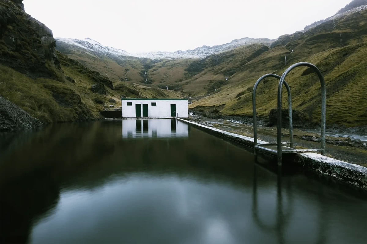 Fotografie budovy šaten venkovních veřejných lázní, izolované uprostřed hornaté islandské přírody, s odrazem v klidné vodní hladině.