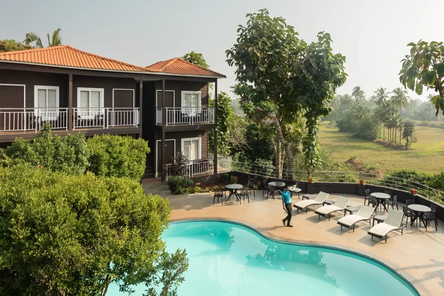 Hotelfotografie eines Resorts: Poolbereich mit Sonnenliegen, umgeben von tropischem Grün und einem zweistöckigen Gebäude mit Balkonen, das auf tropische Felder blickt.