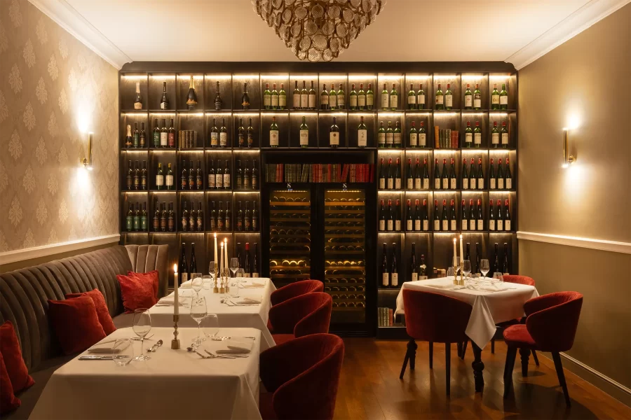 Stylový salonek hotelové restaurace v zámeckém stylu s celostěnovou vinotékou, osvětlený tlumeným ambientním osvětlením a svíčkami, zachycený v čelní kompozici.