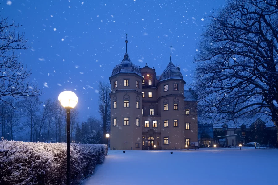 Althörnitz Castle Hotel illuminated at night, surrounded by a serene blanket of snow during snowfall, with a lamp in the foreground.