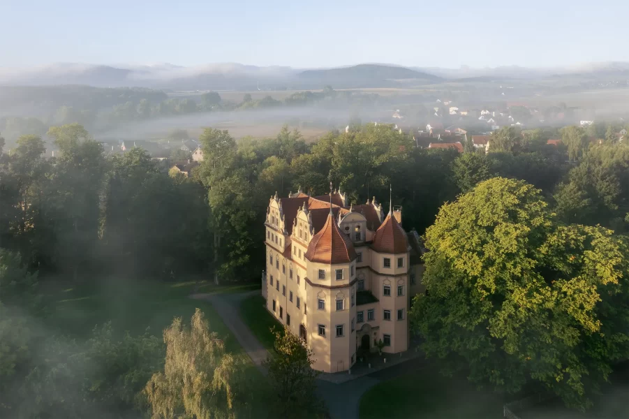 Luftaufnahme des Schlosshotels Althörnitz bei Sonnenaufgang mit Nebel in der umliegenden Landschaft.
