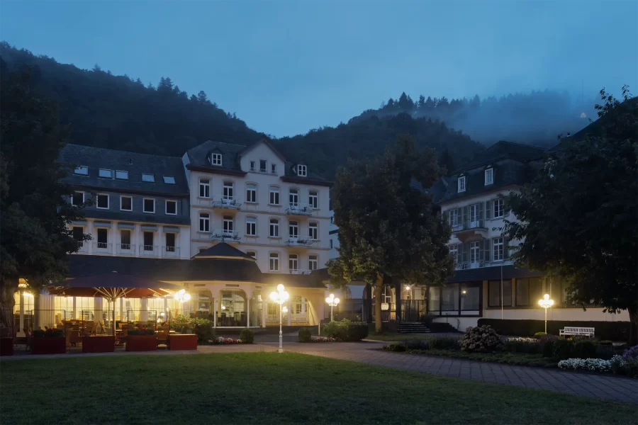 Beleuchtete Caféterrasse des Hotels Fürstenhof in Bad Bertrich zur blauen Stunde, umgeben vom Kurpark und bewaldeten Hügeln.