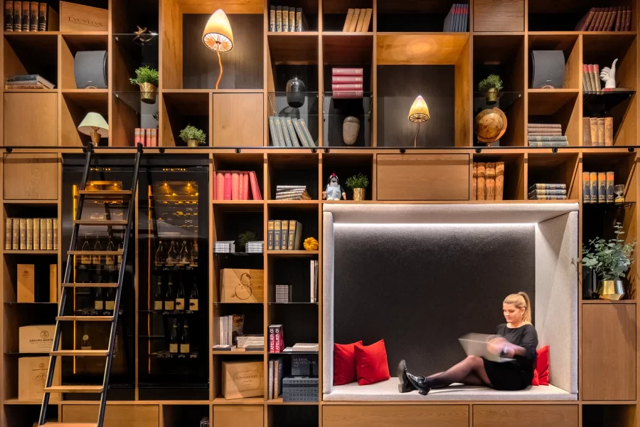 Frontalansicht der Bücherregalwand in der Lobby des Mosaic House in Prag mit integrierter Sitznische, dekorativen Leuchten und einer unscharfen Person bei der Arbeit am Laptop.