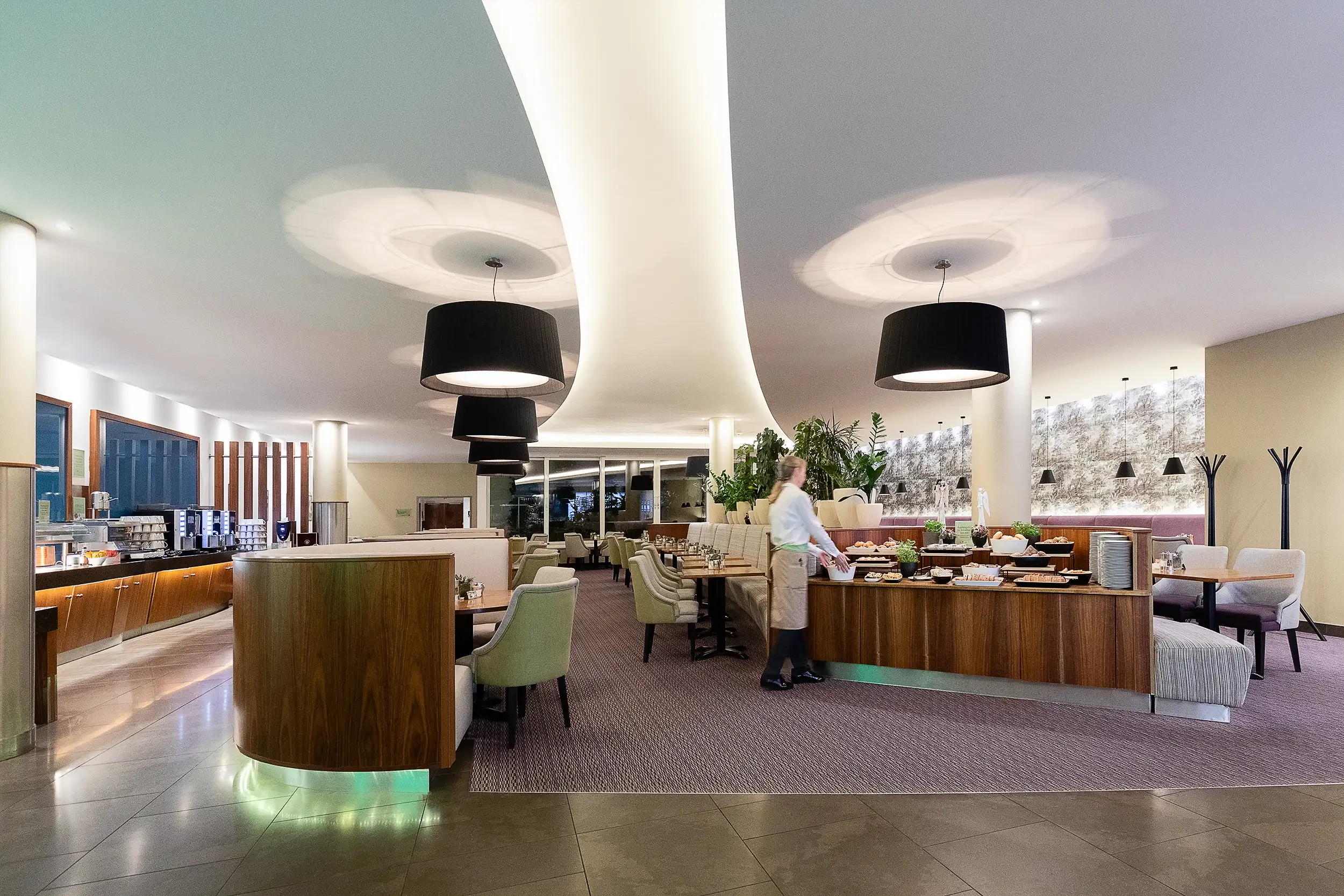 Restaurantbereich mit Frühstücksbuffet, während ein unscharfer Mitarbeiter in Bewegung das Buffet vorbereitet.