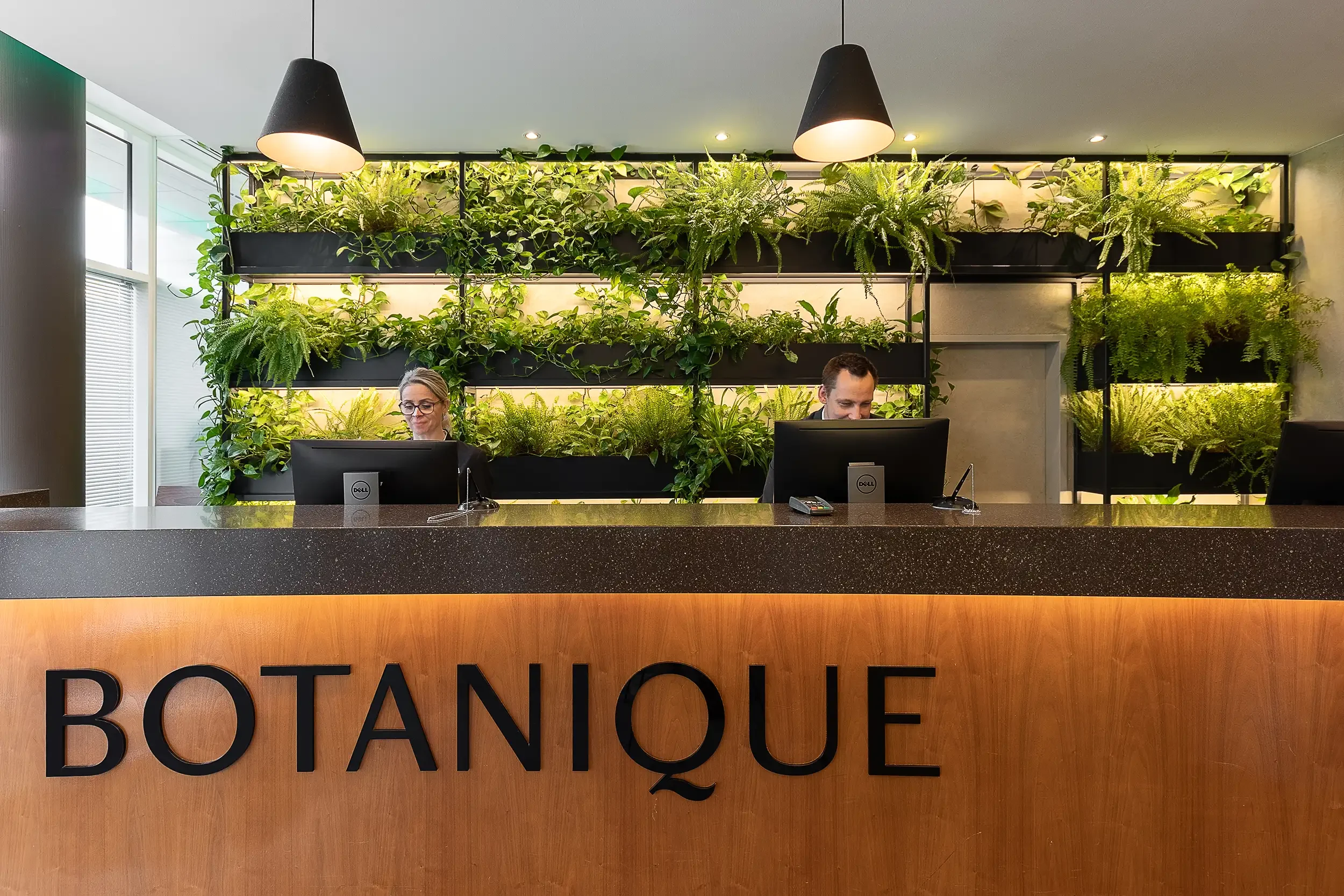 Rezeption im Botanique Prag mit begrüntem Pflanzenregal im Hintergrund und zwei Mitarbeitenden am Empfang.