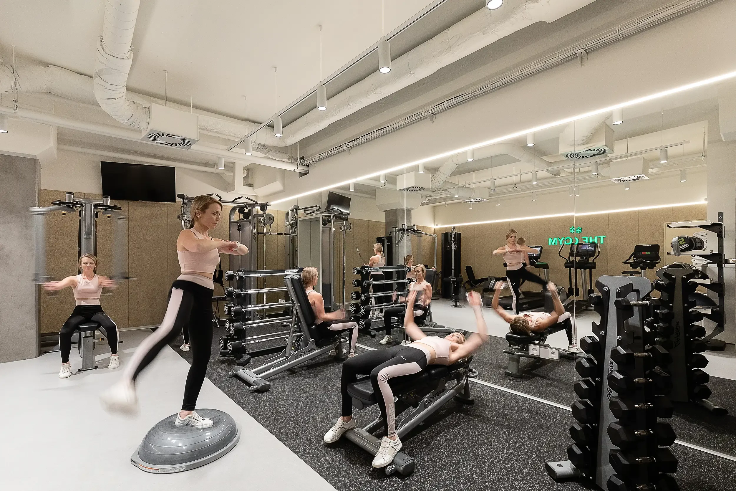 Modernes Fitnessstudio im Botanique Prag mit konzeptioneller Bildidee, bei der dieselbe Frau scheinbar gleichzeitig an mehreren Geräten trainiert.