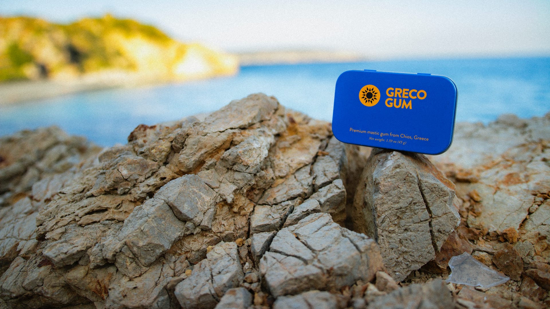 A blue tin of Greco Gum mastic gum nuggets perched atop some cliffs by the ocean in Chios, Greece.