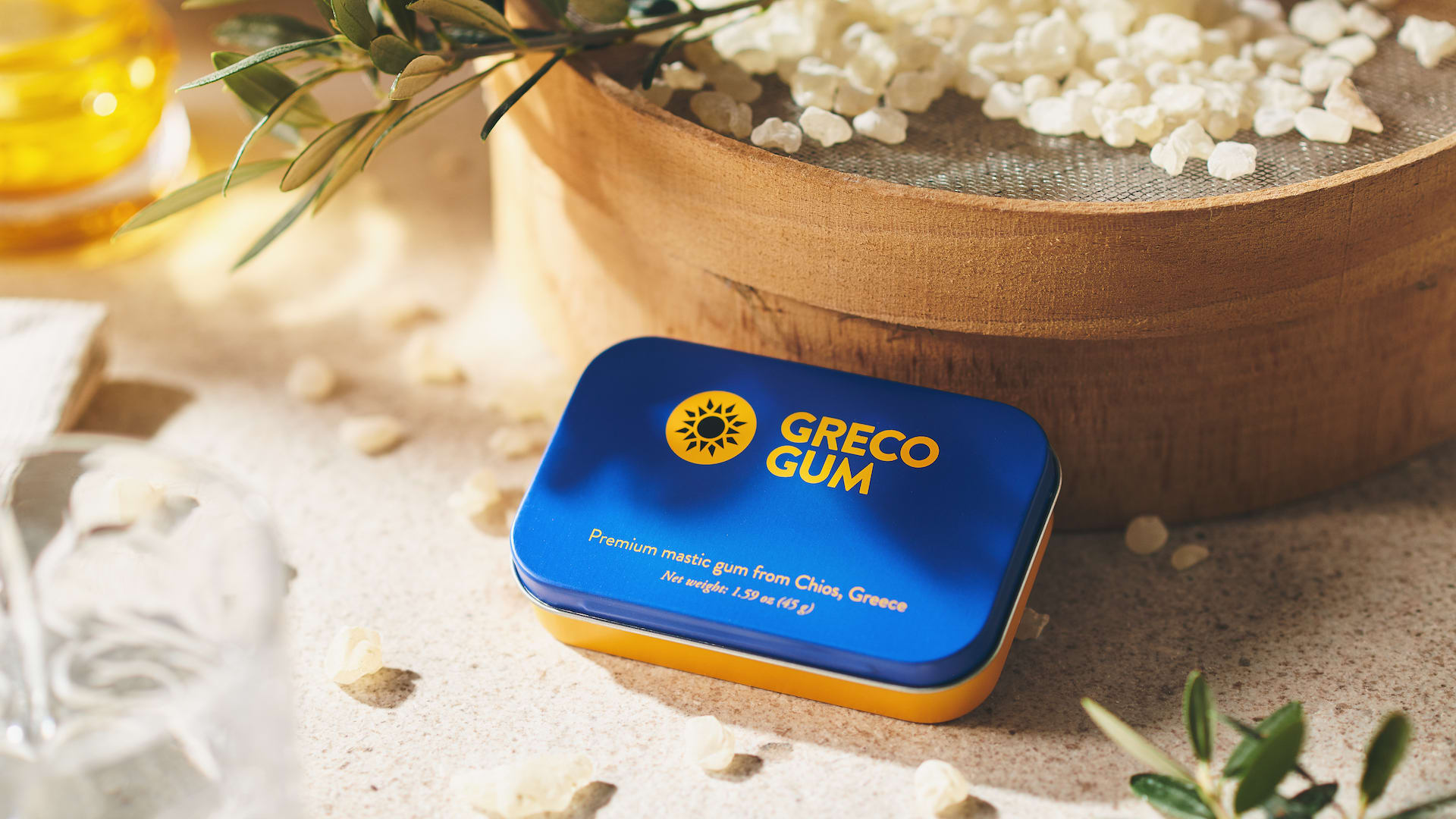 A tin of Greco Gum rests on a table with nuggets of gum sprinkled around it and more nuggets in the background.
